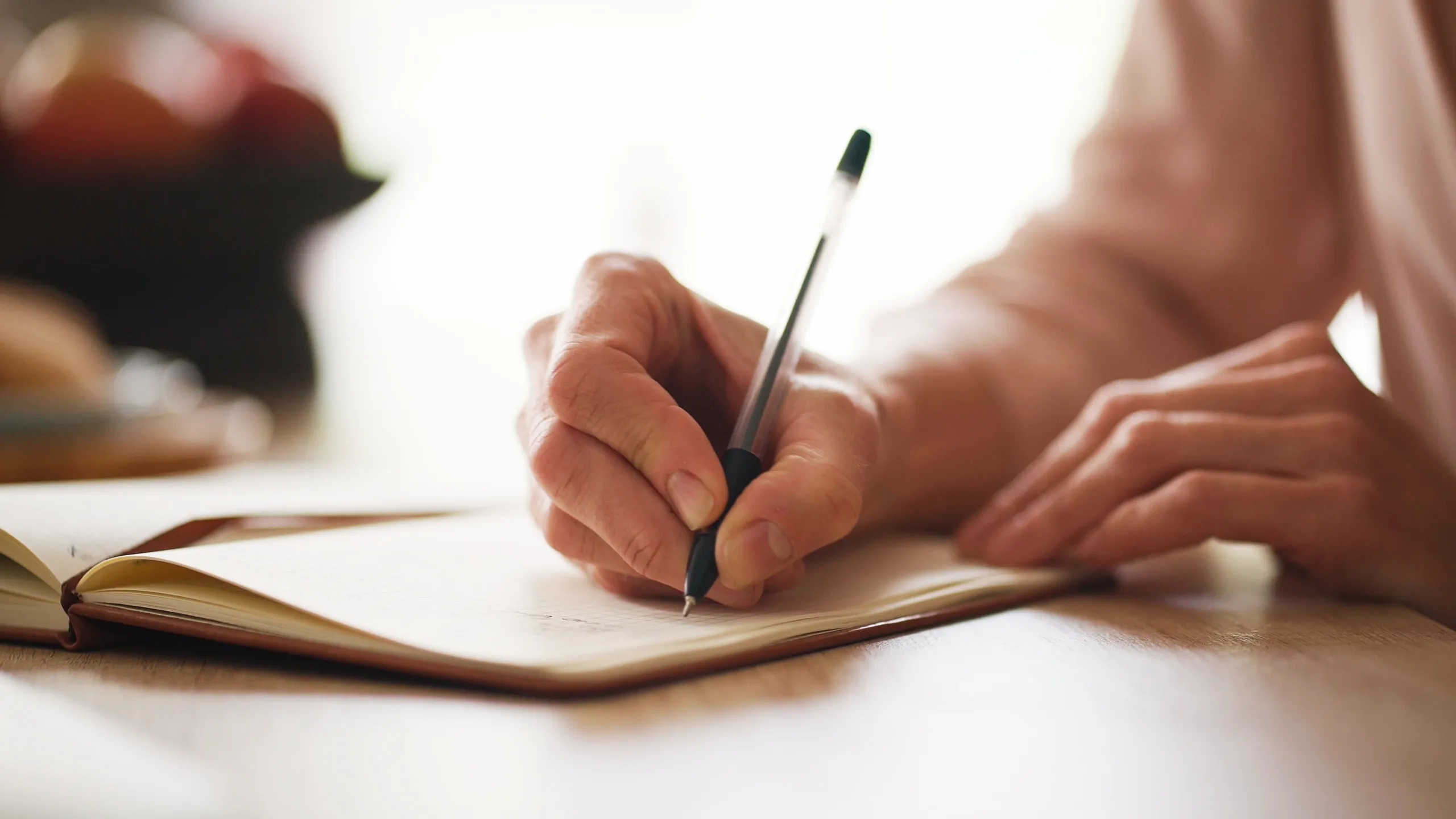 Camera view of woman’s hand holding pen and writing on diary. Female sitting at desk in home or office. Making marks in notepad. Noting shopping list or creating household tasks on paper.